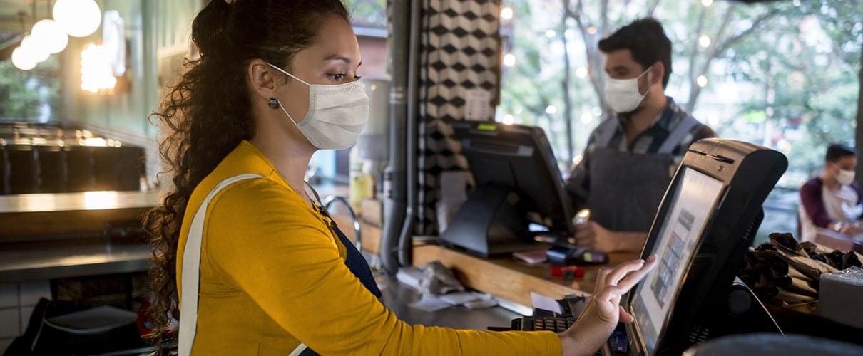 Female restaurant cashier wearing mask using register with male masked employee in background