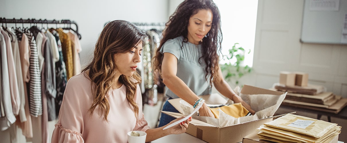 Two women working in a store, drinking coffee and packing online orders and placing in a box