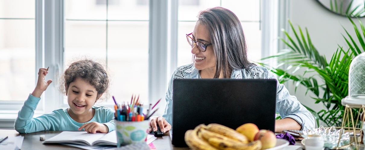 Mother working on laptop sitting next to daughter doing schoolwork