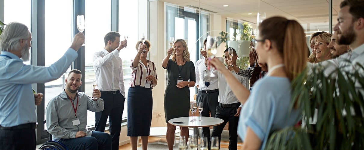 Wide angle view of mature male CEO and diverse business colleagues holding up champagne in preparation for celebratory toast at business event.