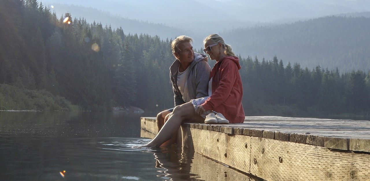 Mature couple sitting on wooden pier with feet in lake and mountains and trees in the background