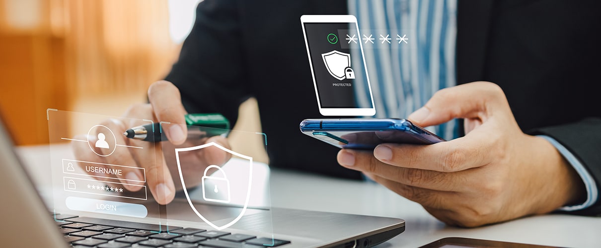 Close-up of a man's hands using a smartphone and laptop with digital security icons illustrated above each device