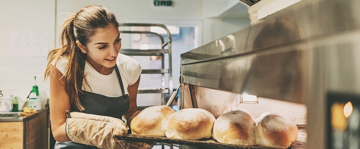 Young woman baker taking out the hot bread from the oven.