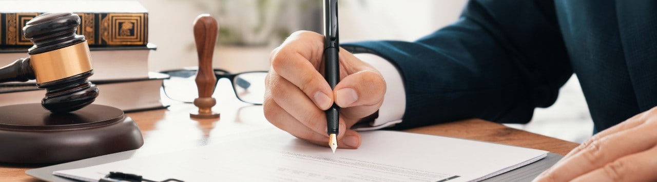 A close-up of masculine hands signing an official document on a desk. A gavel, books, and glasses are on the desk in the background.