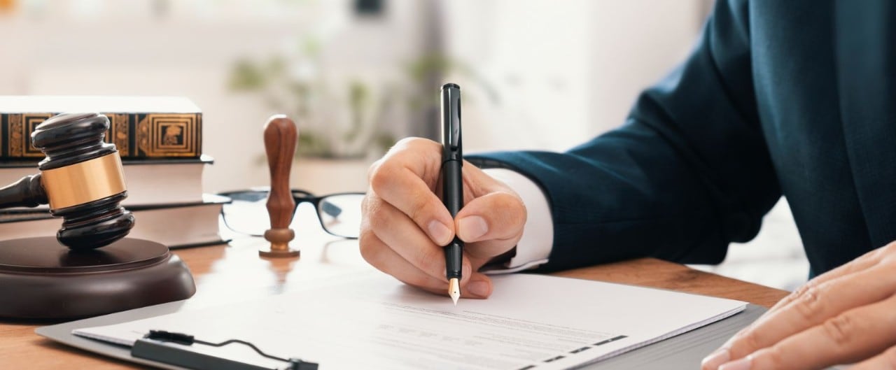 A close-up of masculine hands signing an official document on a desk. A gavel, books, and glasses are on the desk in the background.