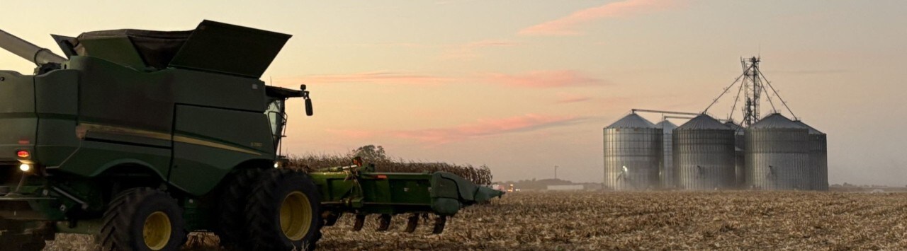 Wide shot of corn field at dusk with combine in foreground and grain elevators in background.