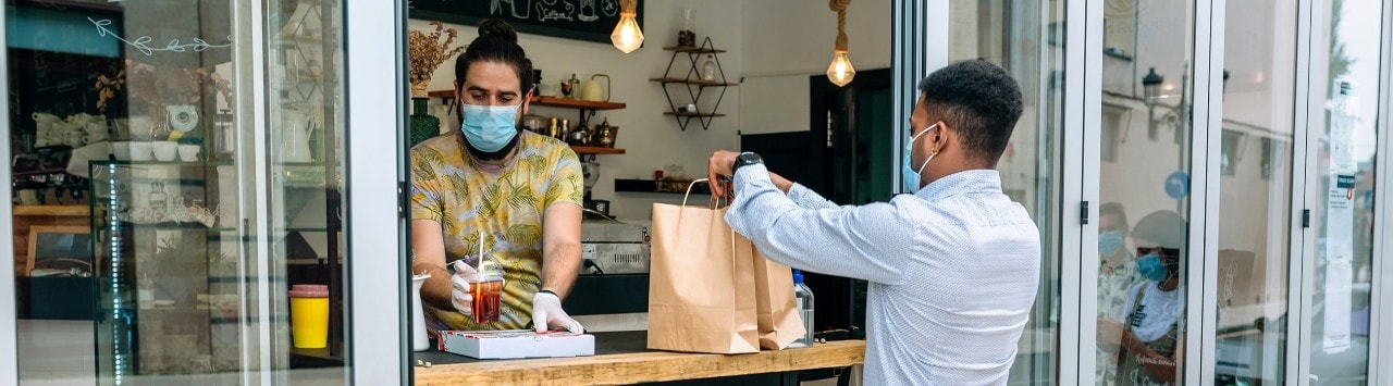 Masked restaurant owner handing order to masked customer through window