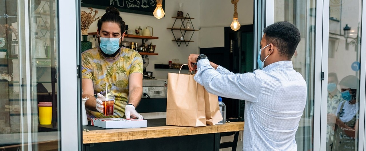 Masked restaurant owner handing order to masked customer through window