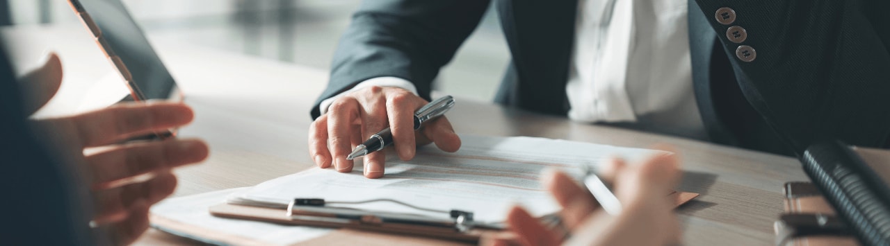 Business professionals review documents during a meeting, with one person holding a tablet and another pointing to paperwork with a pen.