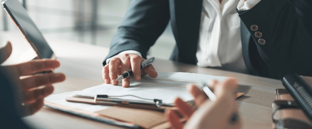 Business professionals review documents during a meeting, with one person holding a tablet and another pointing to paperwork with a pen.