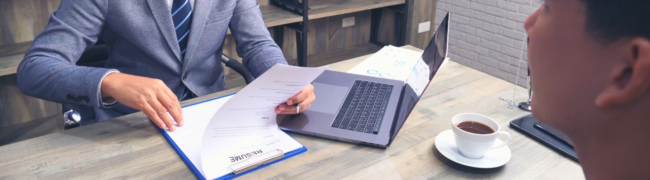Man wearing glasses and a gray suit sits at a desk and reviews a resume while a younger man looks at him from across the desk. 