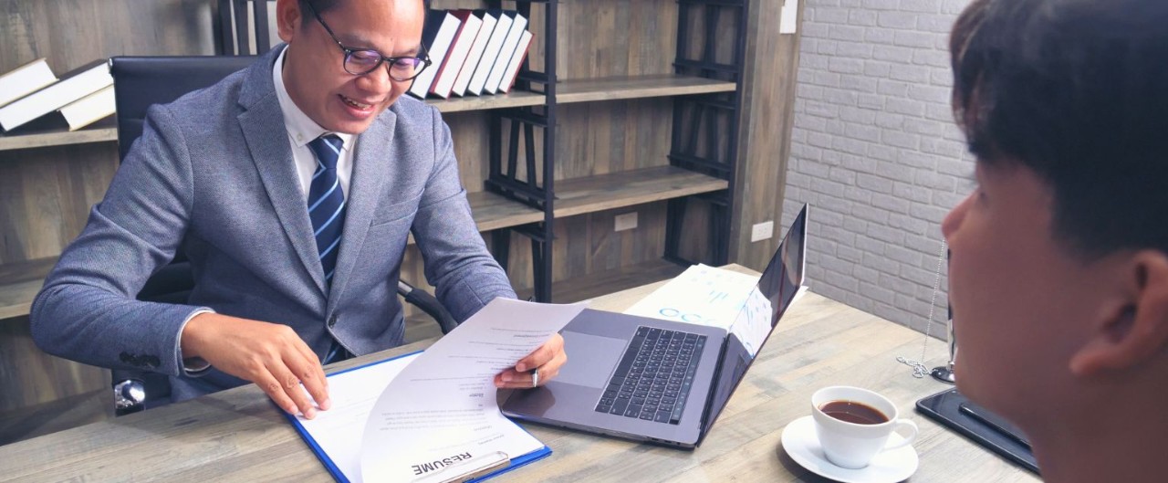 Man wearing glasses and a gray suit sits at a desk and reviews a resume while a younger man looks at him from across the desk. 