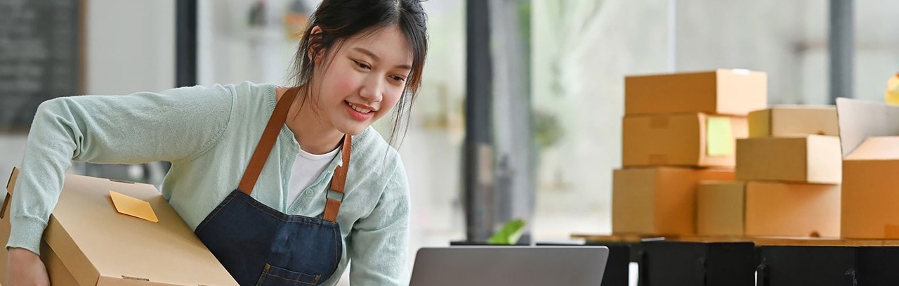 Asian female business owner packing boxes and looking at laptop