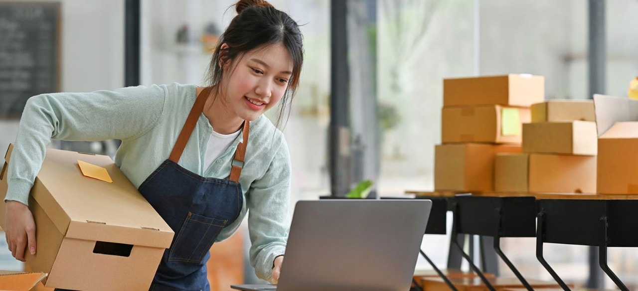 Asian female business owner packing boxes and looking at laptop