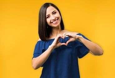 Hispanic woman standing in front of yellow wall with hands forming a heart standing in front of yellow wall with hands forming a heart