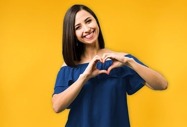 Hispanic woman standing in front of yellow wall with hands forming a heart standing in front of yellow wall with hands forming a heart