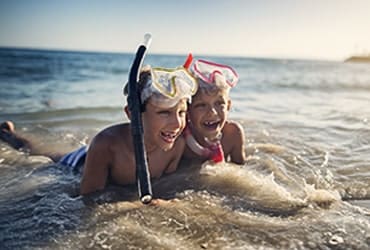 Two young brothers wearing snorkeling masks laying in ocean waves