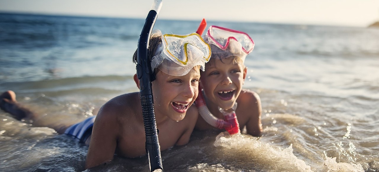Two young brothers wearing snorkeling masks laying in ocean waves