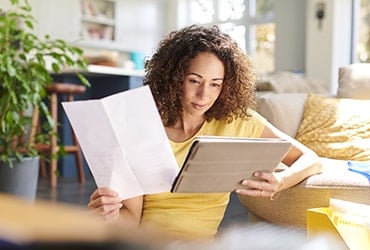 African-American woman leaning against couch holding a piece of paper and a tablet