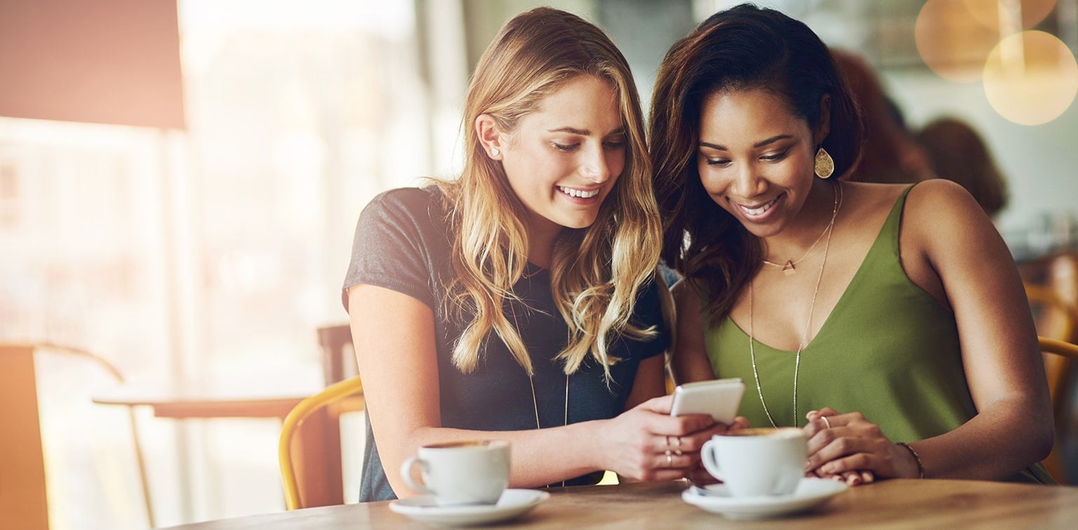 Two young women in coffee shop drinking coffee and looking at smartphone