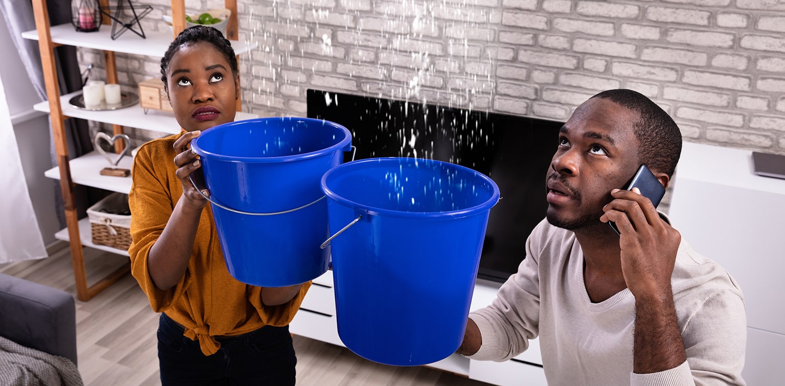 Couple using buckets to collect water from leaky ceiling while calling plumber