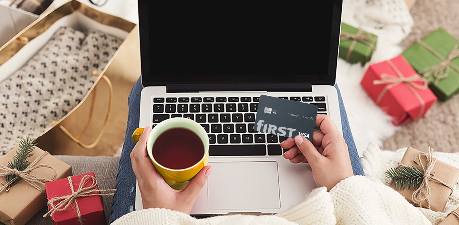 Woman holding First Financial Rewards card and a cup of coffee looking at laptop surrounded by holiday gifts