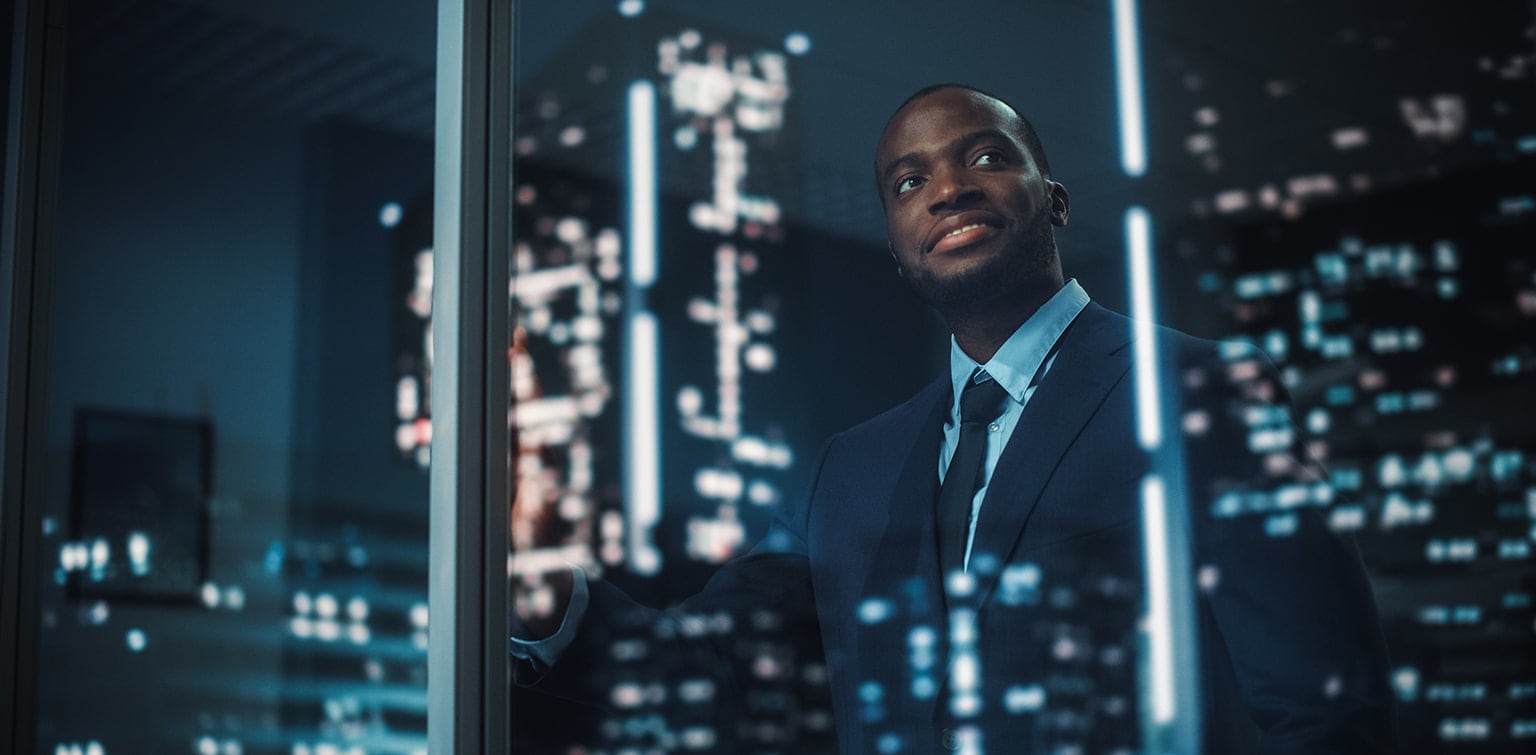 Happy, young African-American businessman looking out window at city at night