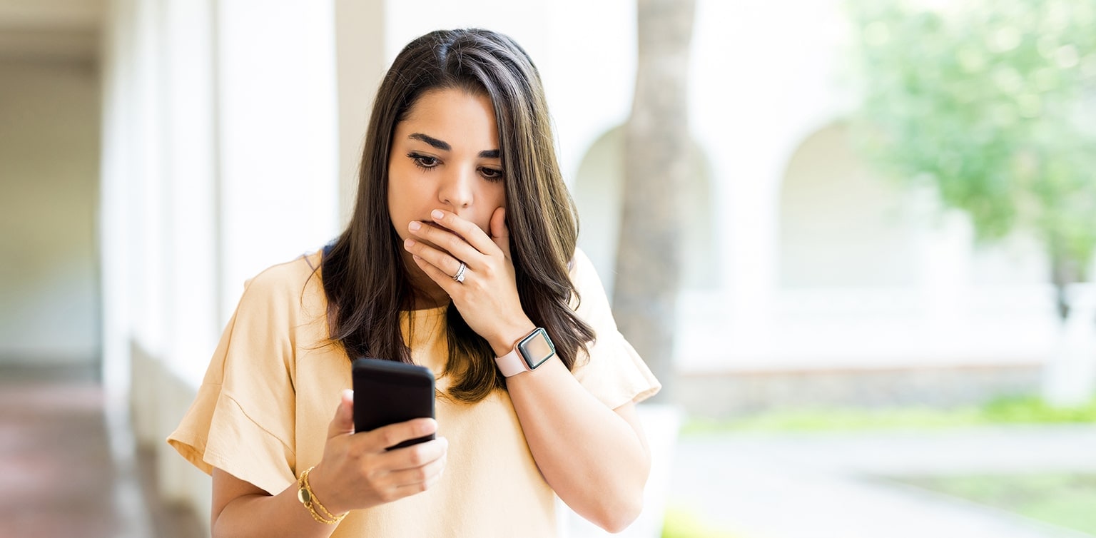Young woman holding her hand over mouth looking at her smartphone with a shocked look