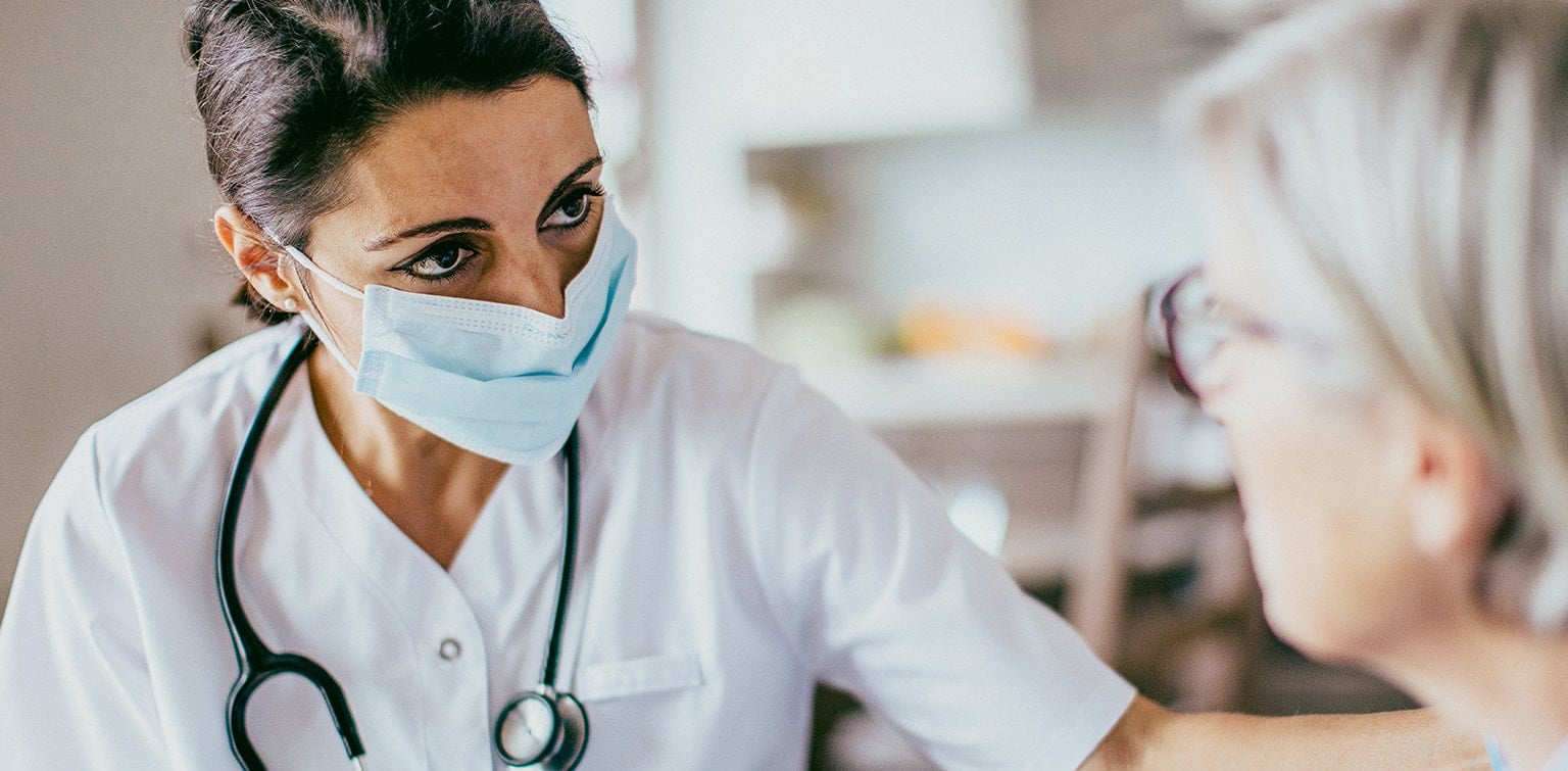 A medical professional in scrubs in mask with a stethoscope around her neck observing a patient.