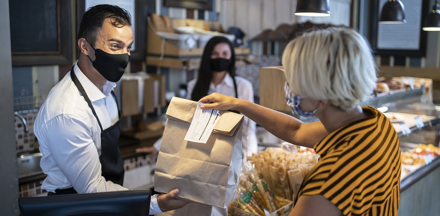 Masked deli employee handing carryout bag to customer