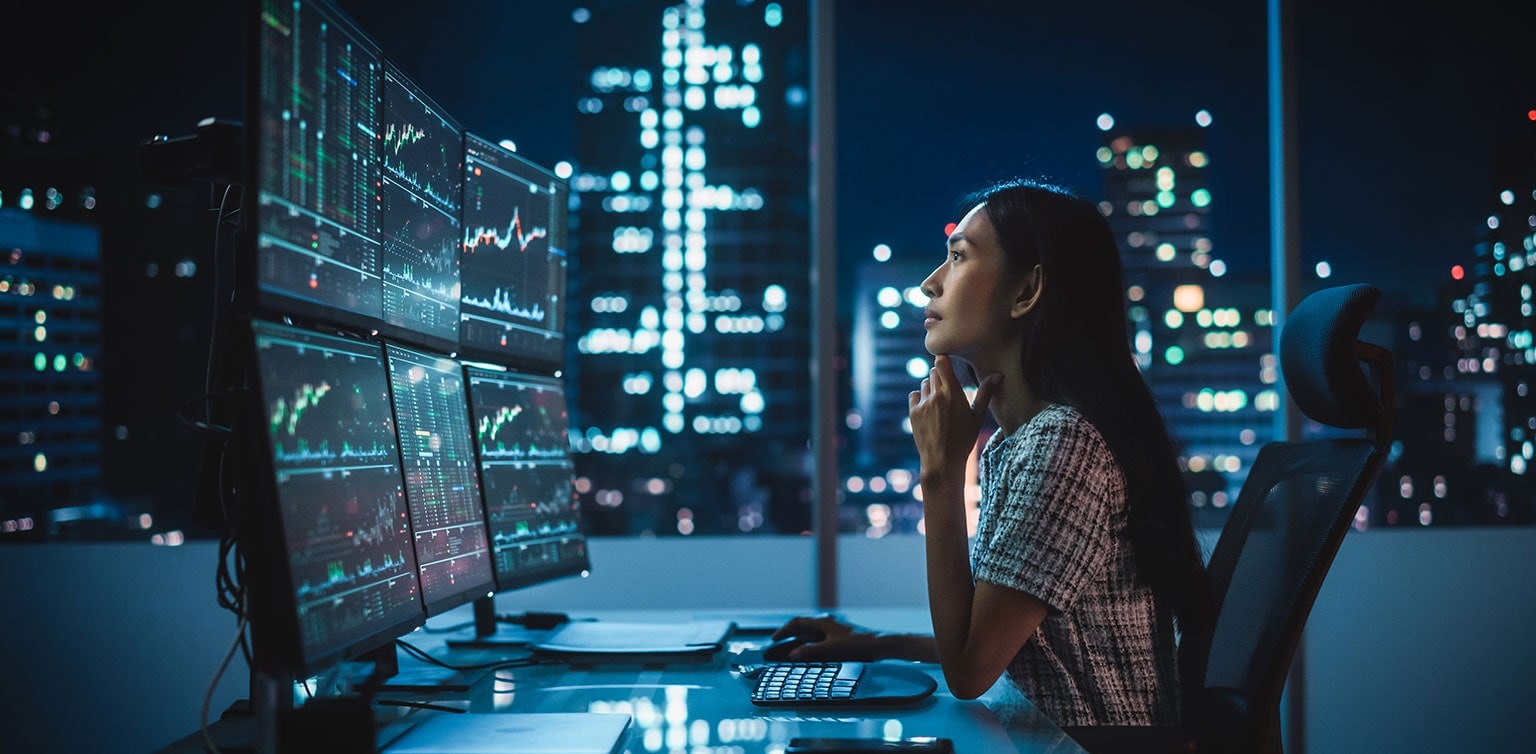 Asian female financial analyst reviewing stock data on multiple monitors