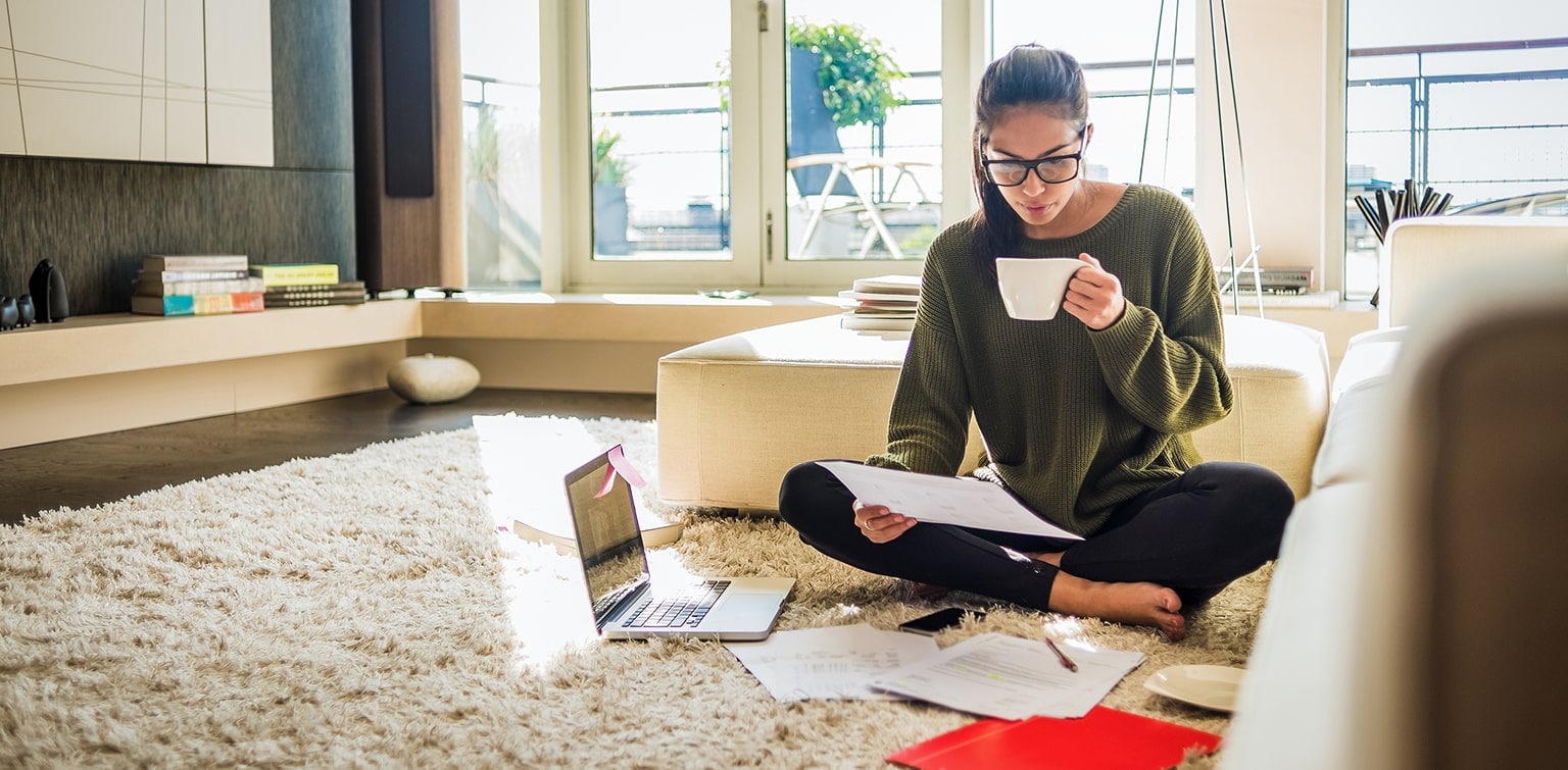 Young college grad sitting on floor at home reviewing bills and drinking coffee