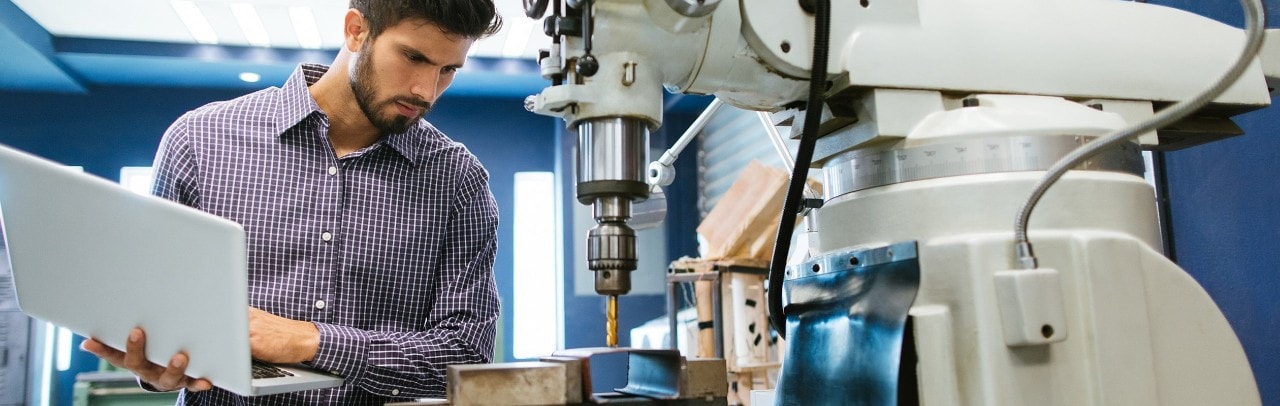 A man works on a laptop in front of a large industrial machine.