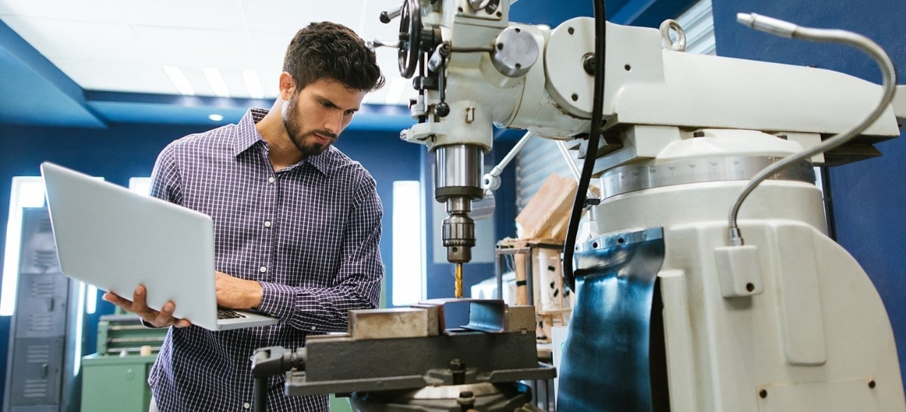 A man works on a laptop in front of a large industrial machine.