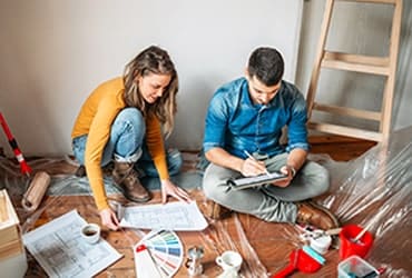 Young couple sitting on floor during home renovation looking at blueprint and table