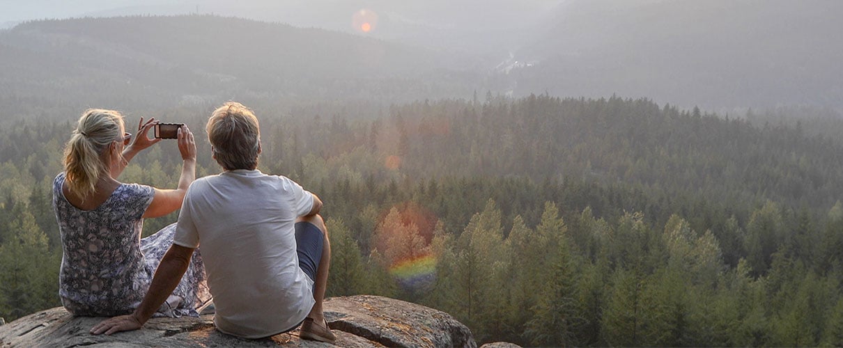 A couple seated on a cliff taking a photo of a mountain landscape