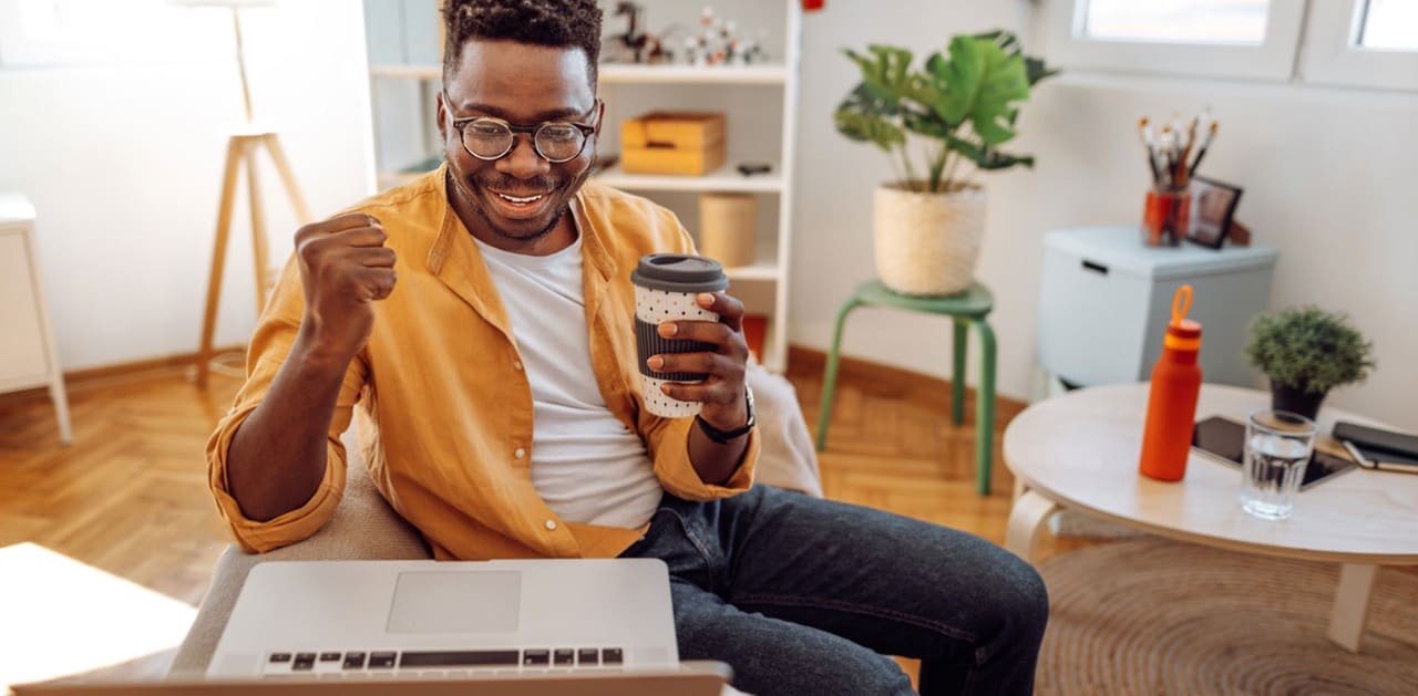 Cheerful African-American male college student holding coffee and looking at laptop