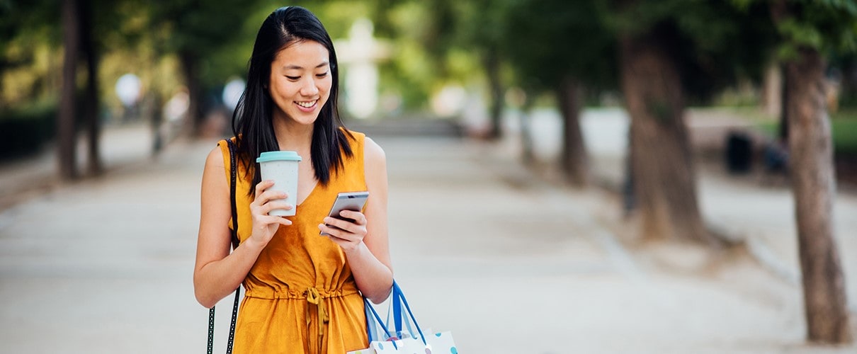Young Asian woman holding coffee cup and shopping bags and looking at smartphone