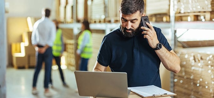 Man talking on smartphone and typing on laptop in a warehouse with coworkers in the background