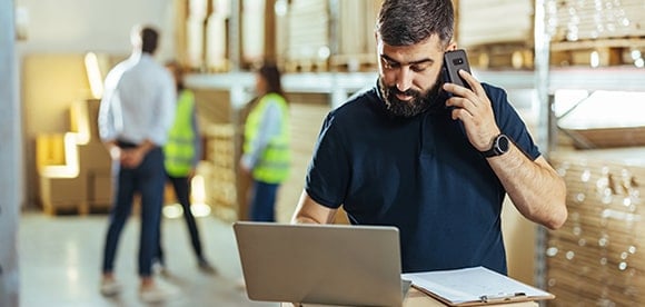 Man talking on smartphone and typing on laptop in a warehouse with coworkers in the background