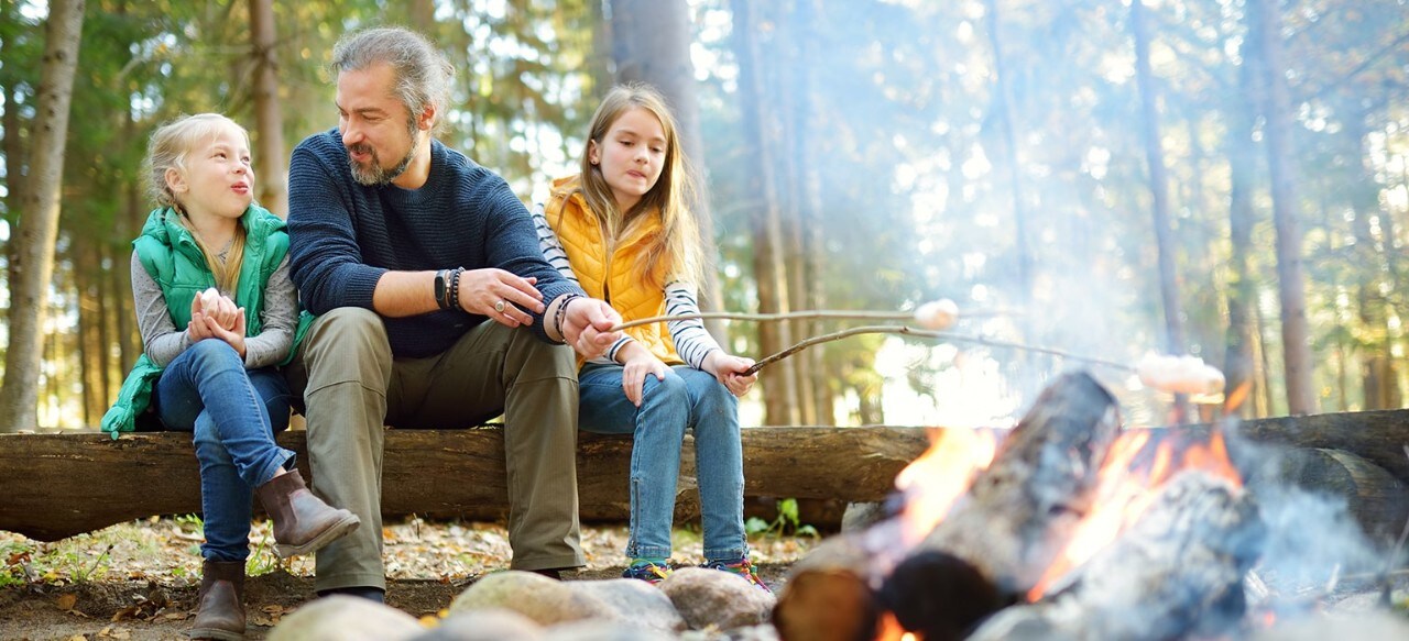 Father at campsite with two young daughters roasting marshmallows in fire pit
