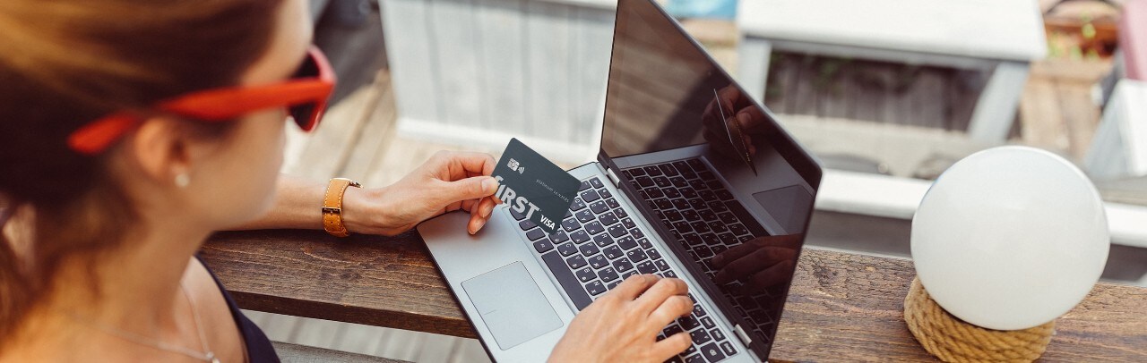 Woman wearing sunglasses at outdoor bar using laptop holding f1RST® Platinum rewards card