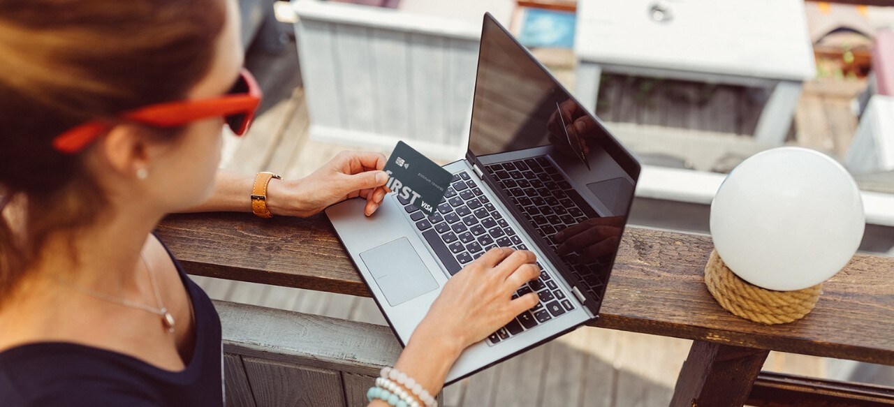 Woman wearing sunglasses at outdoor bar using laptop holding f1RST® Platinum rewards card