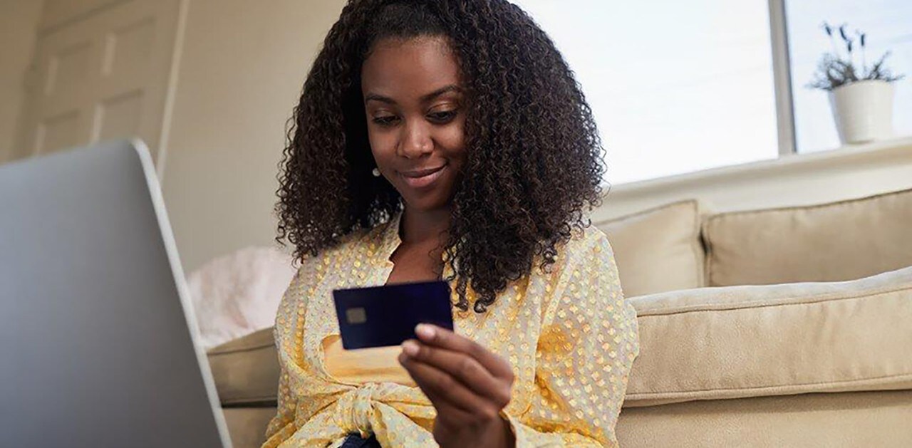 African-American woman leaning against couch holding credit card and laptop