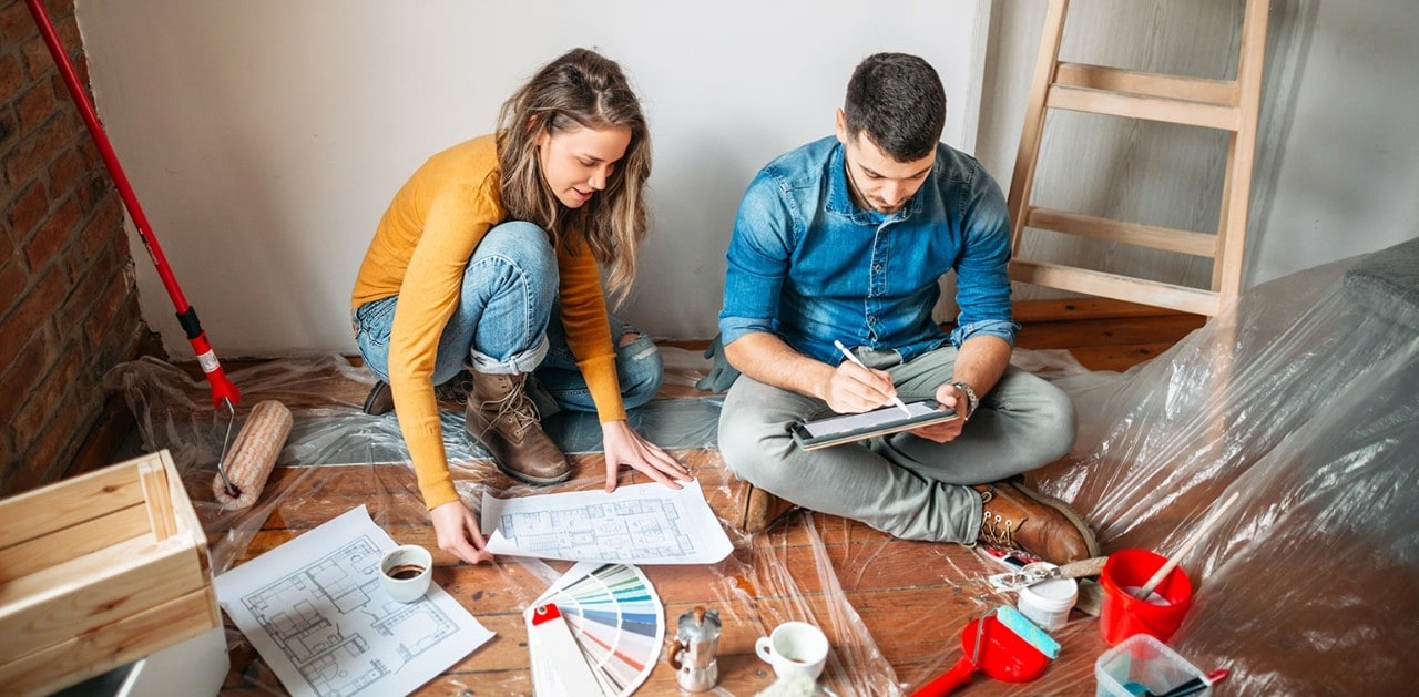 Young couple sitting on floor during home renovation looking at blueprint and table