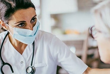 A medical professional in scrubs in mask with a stethoscope around her neck observing a patient.