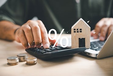Close-up of a man's hands using a calculator and laptop, with coins and wooden house model nearby