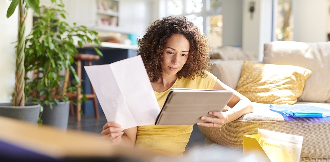 Woman leaning on couch holding paper and looking at tablet