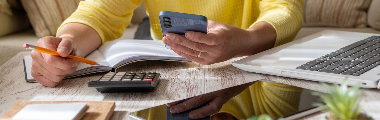 A woman holding a pencil and smartphone while using a calculator, laptop, and tablet to review documents