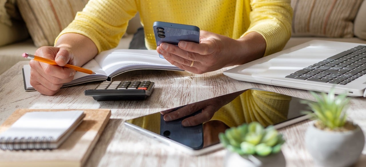 A woman holding a pencil and smartphone while using a calculator, laptop, and tablet to review documents